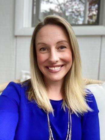 A woman with shoulder-length blonde hair and a bright smile, wearing a blue top and a necklace, seated indoors with a window in the background, ready to guide college students on how to manage stress.