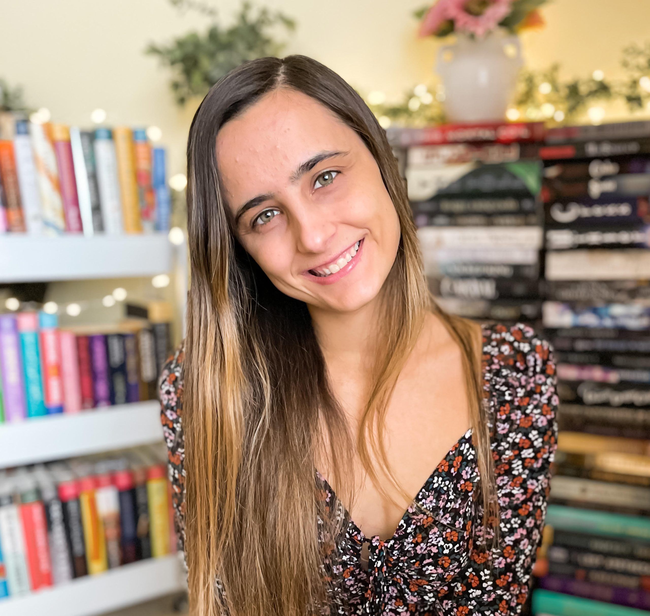A smiling woman with long brown hair in a floral dress stands in front of a bookshelf filled with colorful books, with string lights and a vase of flowers in the background.