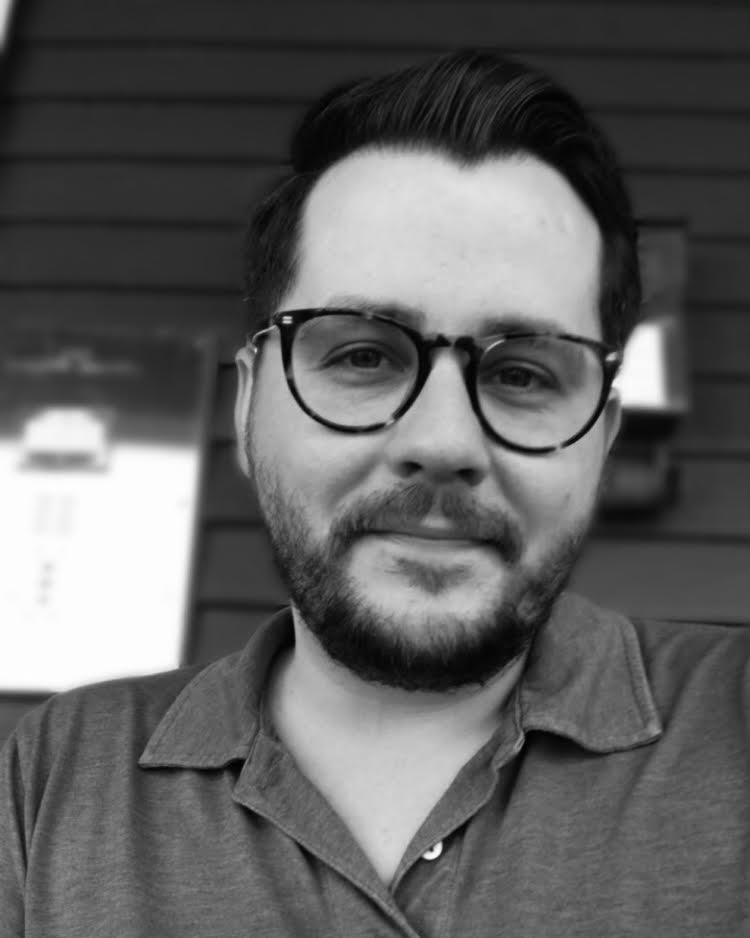 A black and white portrait of a man with glasses, short beard, and moustache, wearing a polo shirt, slightly smiling at the camera.