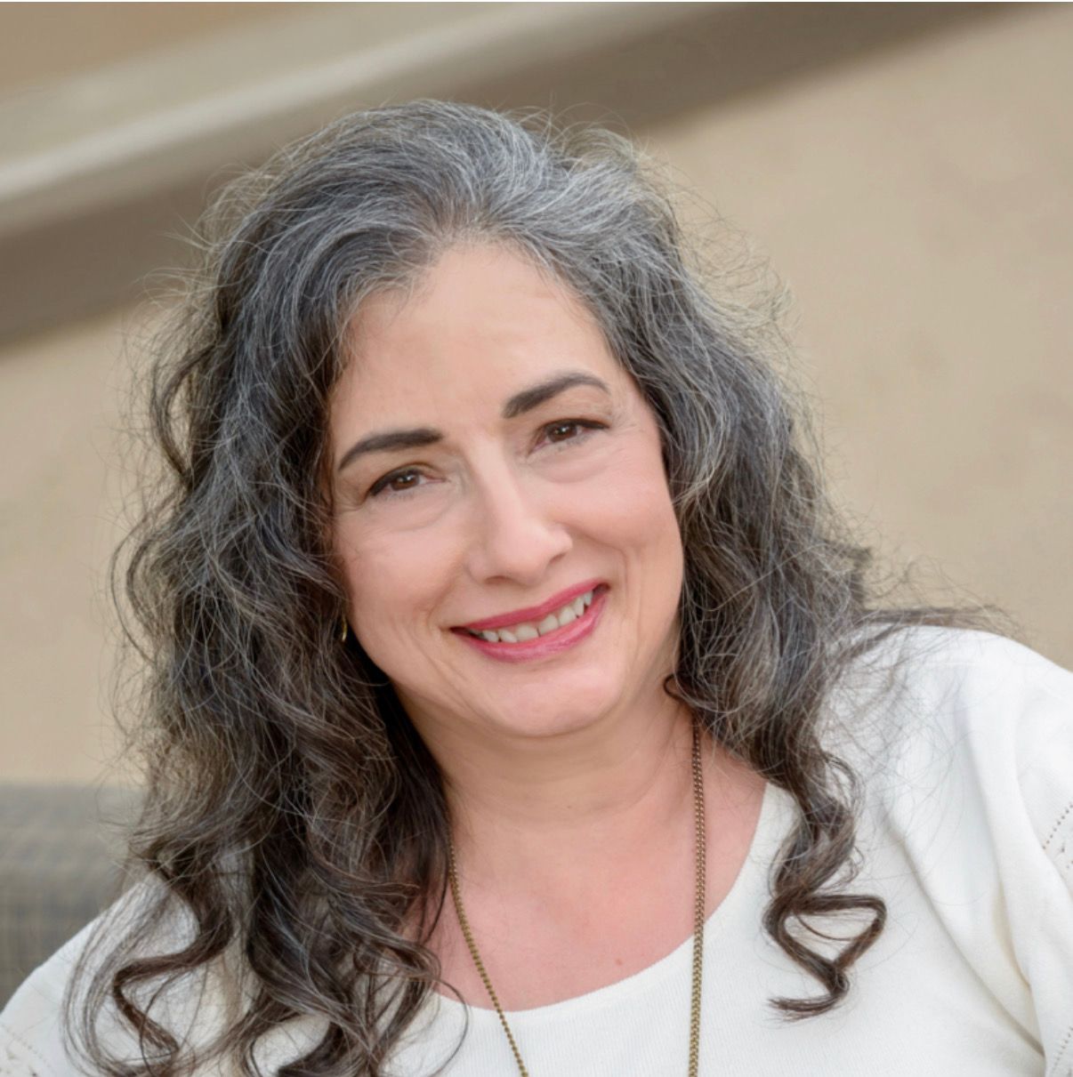 A portrait of a smiling woman with shoulder-length curly gray hair, wearing a light beige top. She appears outdoors with a soft-focus background.
