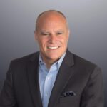 Professional headshot of a smiling middle-aged man wearing a dark blazer, light blue shirt, and pocket square against a gray background.