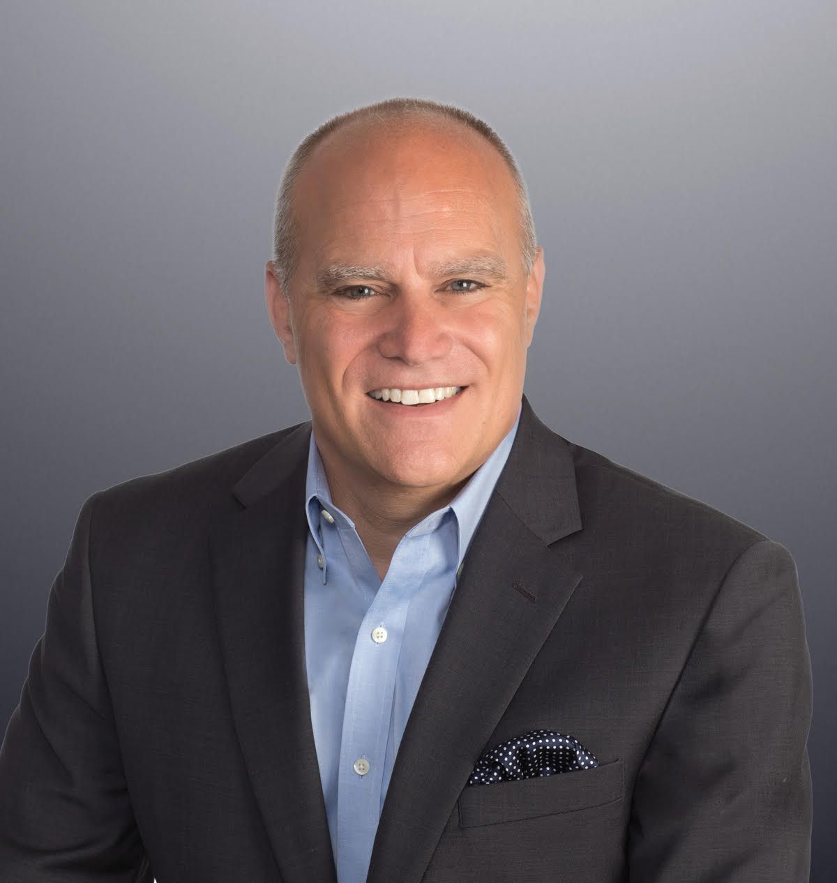 Professional headshot of a smiling middle-aged man wearing a dark blazer, light blue shirt, and pocket square against a gray background.