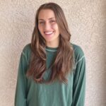 A smiling young woman with long brown hair wearing a green sweater, standing against a textured beige wall.