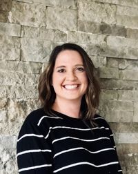 A woman with shoulder-length hair smiling at the camera, wearing a black and white striped top. She stands in front of a textured stone wall.