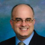 Professional headshot of a smiling bald man wearing glasses and a dark suit with a patterned tie against a blue gradient background.