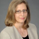 A professional headshot of a woman with shoulder-length blond hair and glasses, wearing a beige blazer over a black top, with a neutral background.