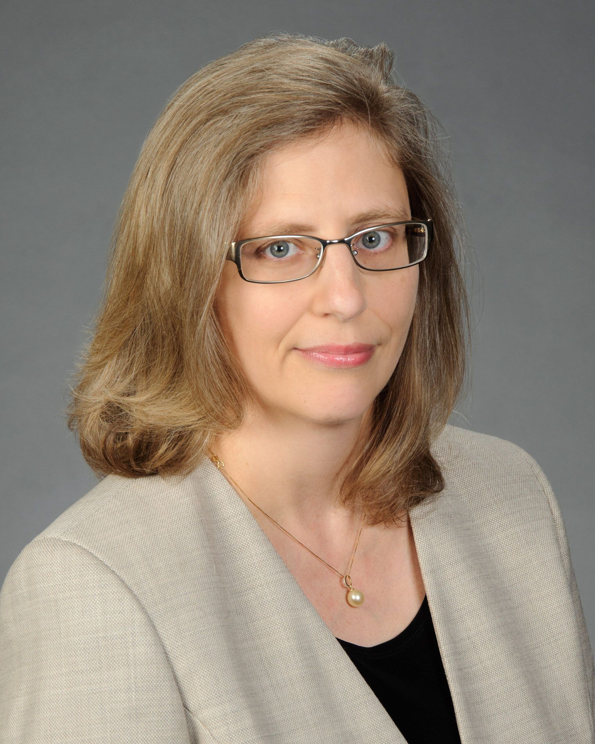 A professional headshot of a woman with shoulder-length blond hair and glasses, wearing a beige blazer over a black top, with a neutral background.