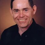 A portrait of a smiling middle-aged man with short dark hair, wearing a plain black button-up shirt against a soft-focused maroon background.