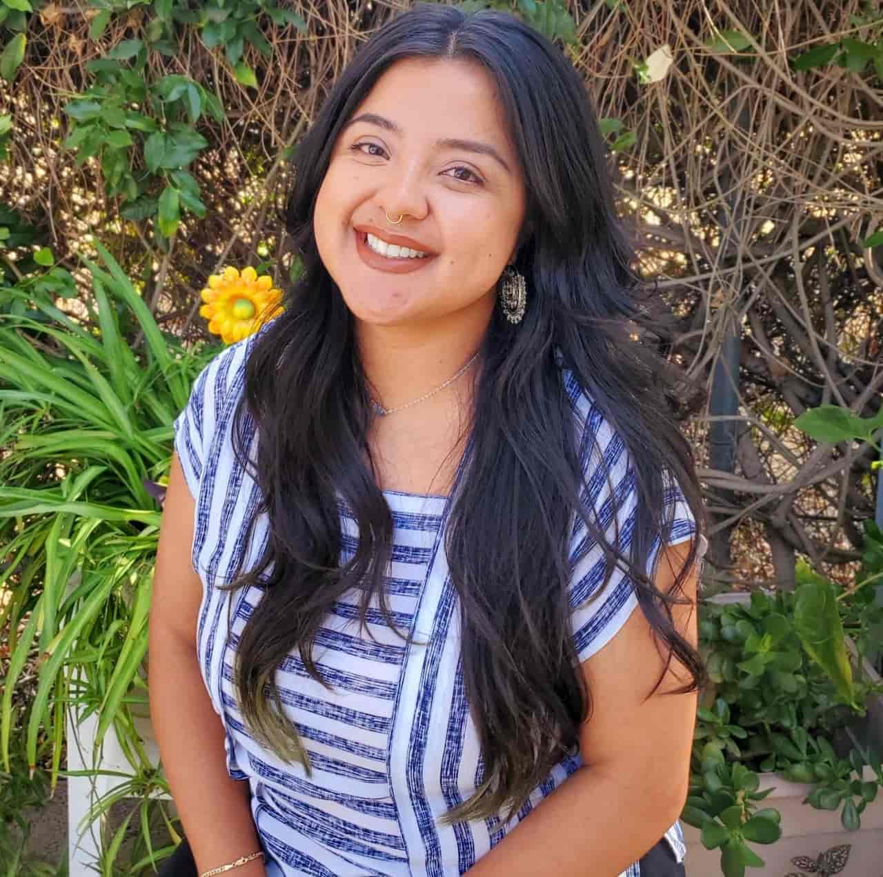 A smiling woman with long dark hair wearing a striped blue and white top, standing in a garden with plants and a yellow flower in the background.