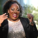A joyful woman in a sparkling top and black blazer laughs outdoors, adjusting her earring with a backdrop of lush greenery.