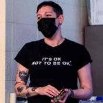 A woman wearing a black mask and a T-shirt that reads "It's OK NOT TO BE OK," stands while holding cards, with visible tattoos on her arms.