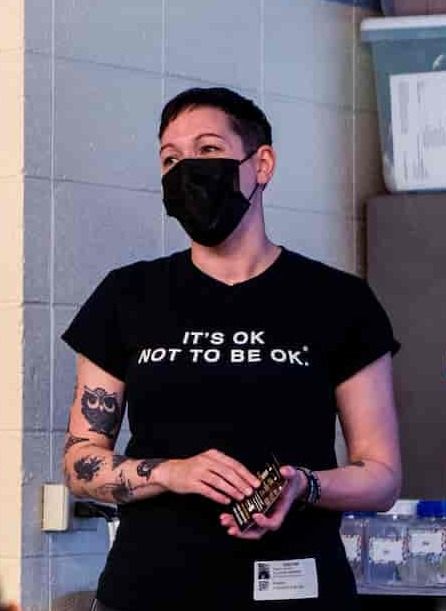 A woman wearing a black mask and a T-shirt that reads "It's OK NOT TO BE OK," stands while holding cards, with visible tattoos on her arms.
