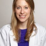 A professional portrait of a smiling woman with long blonde hair, wearing a white lab coat and a blue top, against a light gray background.