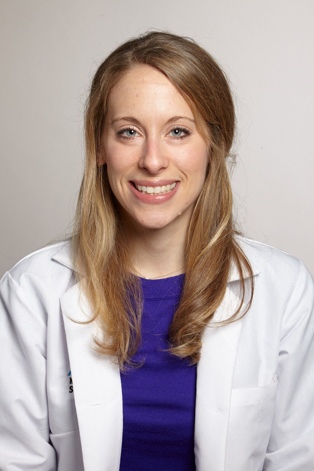 A professional portrait of a smiling woman with long blonde hair, wearing a white lab coat and a blue top, against a light gray background.