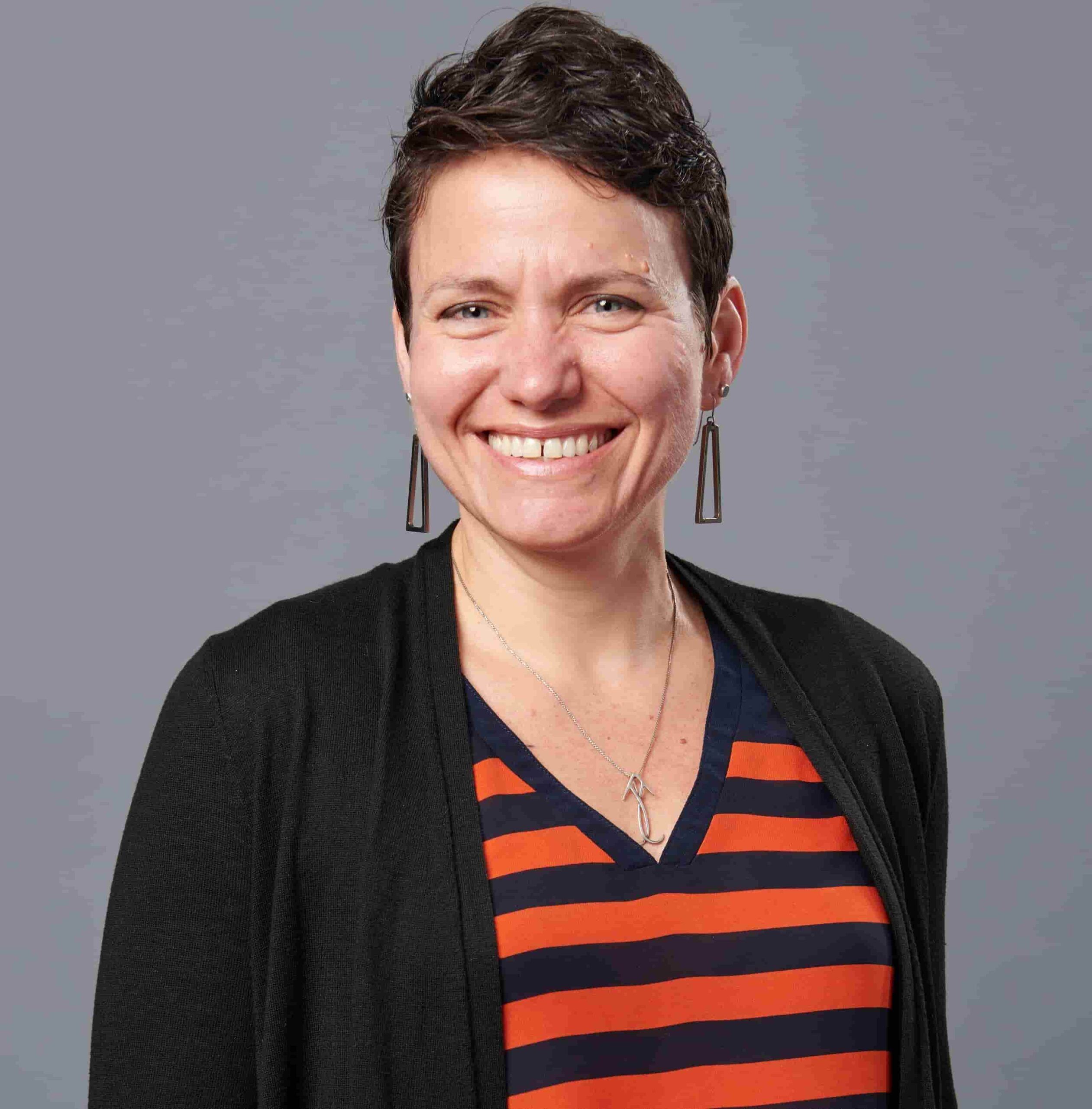 A smiling woman with short brown hair and earrings, wearing a black cardigan over a striped top, standing against a gray background.