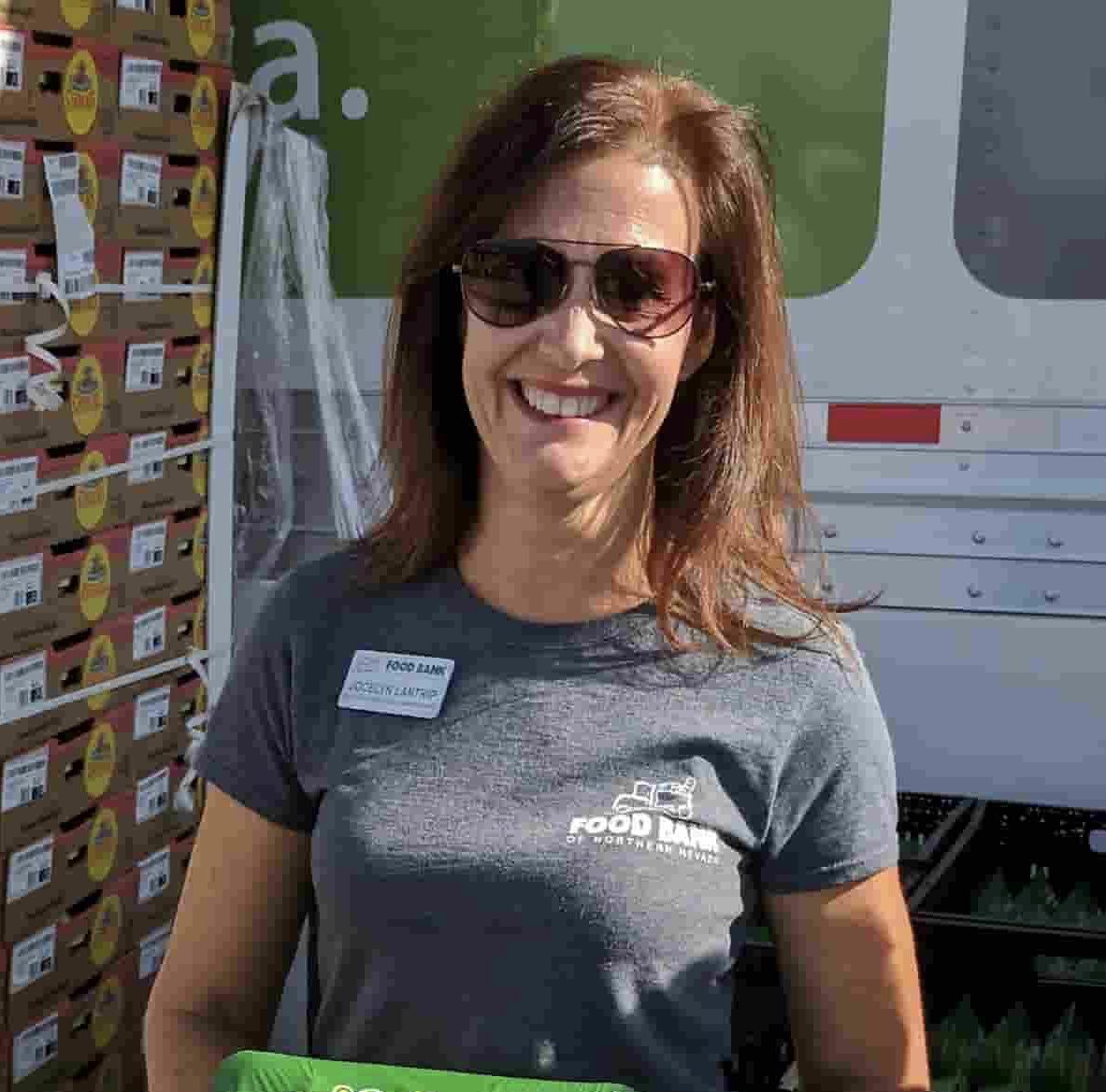 A smiling woman wearing sunglasses and a gray T-shirt with "Food Bank" and a logo on it stands in front of a truck loaded with boxes of food donations.