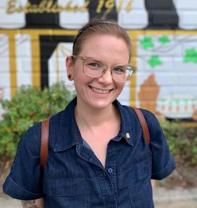 A smiling woman with glasses, wearing a denim shirt and a shoulder bag, stands in front of a mural with the text "Established 1916.