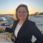 A woman smiling, with a marina and sunset in the background. She is wearing a dark blazer and standing on a balcony.
