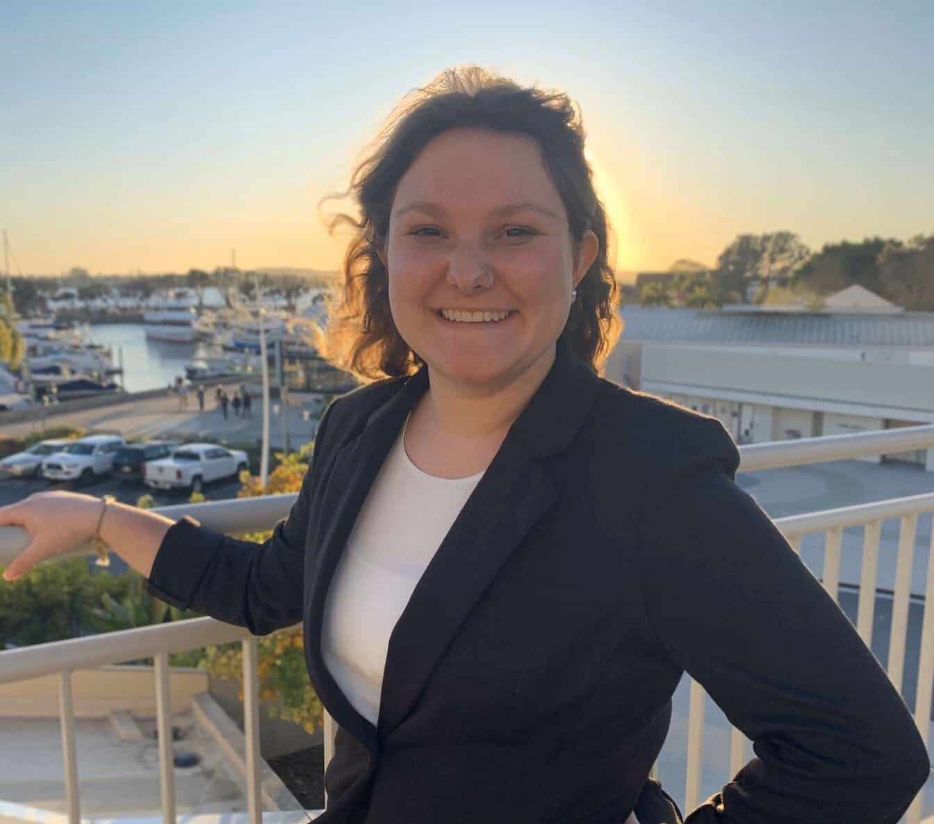A woman smiling, with a marina and sunset in the background. She is wearing a dark blazer and standing on a balcony.