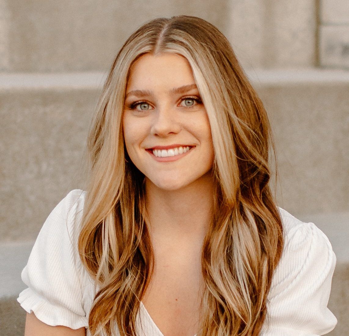 A smiling young woman with long wavy blonde hair, wearing a white top, posing outdoors with a soft-focus background.