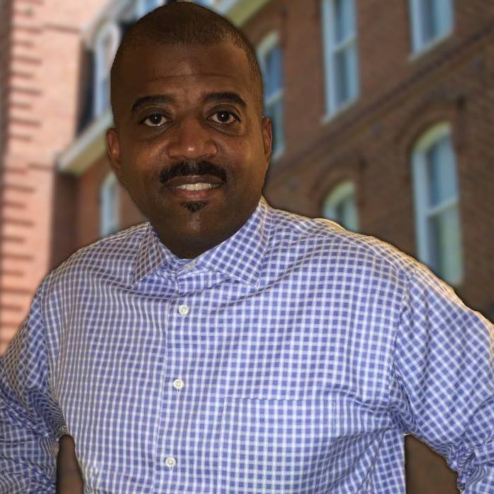 A middle-aged man with a mustache smiles at the camera, wearing a blue checked shirt. He is posed against a blurred backdrop of brick buildings.