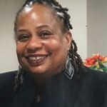 A smiling woman with braided hair and large earrings, wearing a black outfit. She is in a room with a blurred background showing orange flowers.