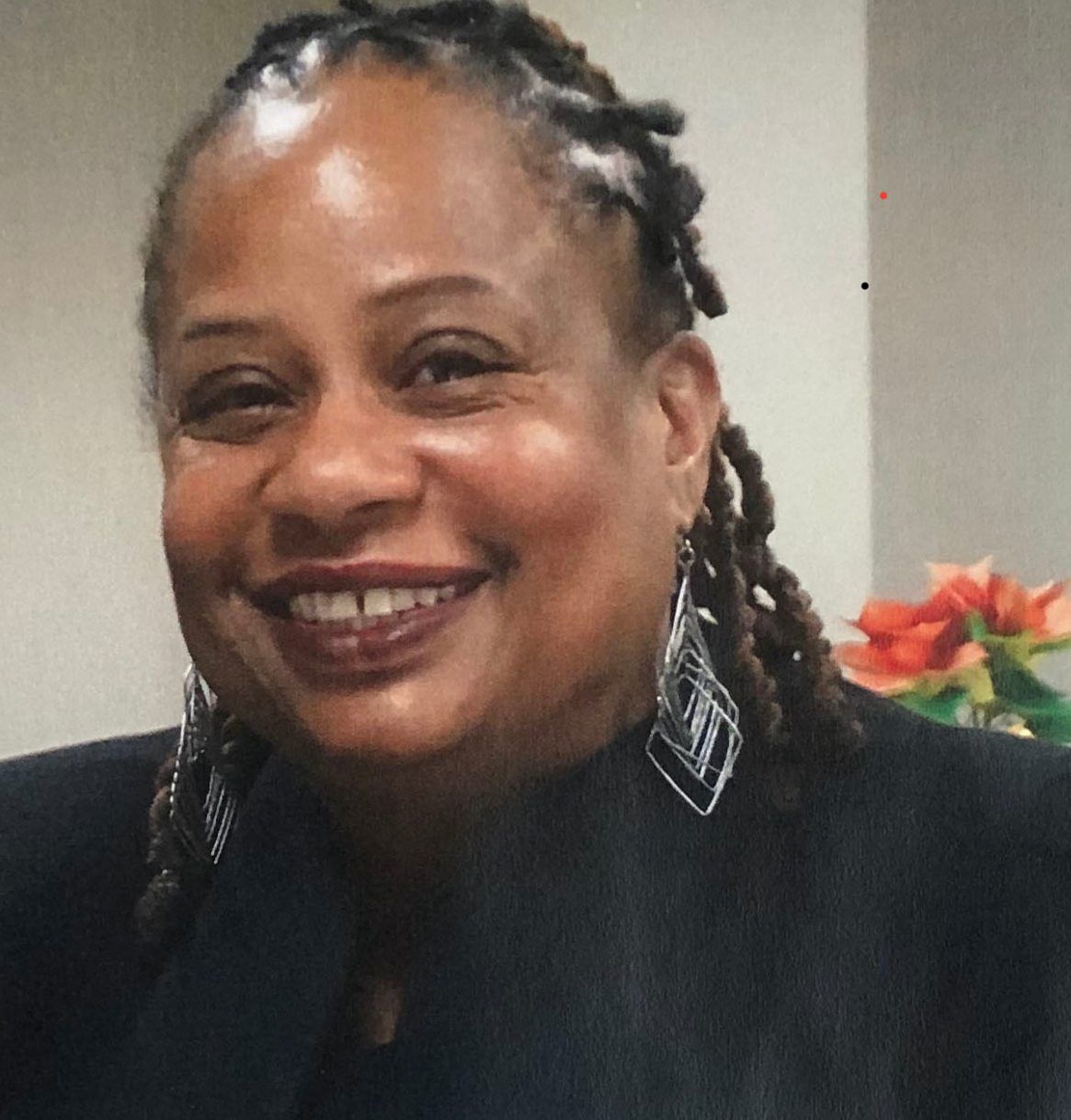 A smiling woman with braided hair and large earrings, wearing a black outfit. She is in a room with a blurred background showing orange flowers.