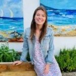 A woman with long brown hair in a floral dress and denim jacket, smiling and sitting on a wooden bench against a backdrop of vibrant ocean paintings.