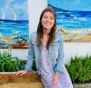 A woman with long brown hair in a floral dress and denim jacket, smiling and sitting on a wooden bench against a backdrop of vibrant ocean paintings.