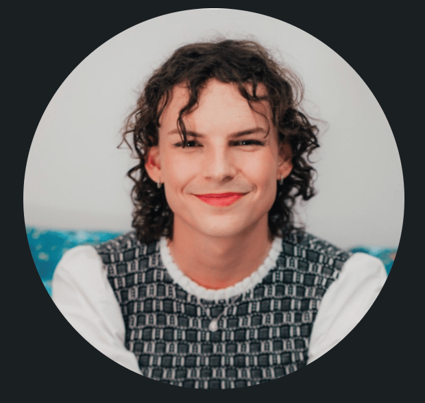 A portrait of a person with shoulder-length curly hair, smiling and wearing a white blouse under a checkered vest, against a blurred indoor background.