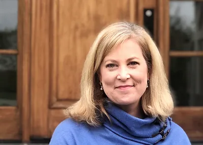 A smiling woman with blonde hair, wearing a blue scarf, stands in front of a wooden door.