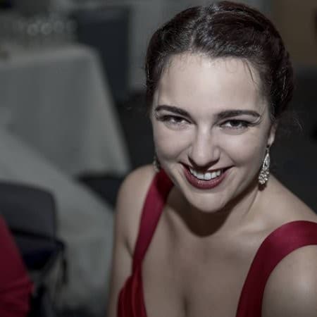 A woman with dark hair in an elegant red dress smiles at the camera, her face lit warmly in a dimly lit room. She wears earrings and her makeup accentuates her cheerful expression.