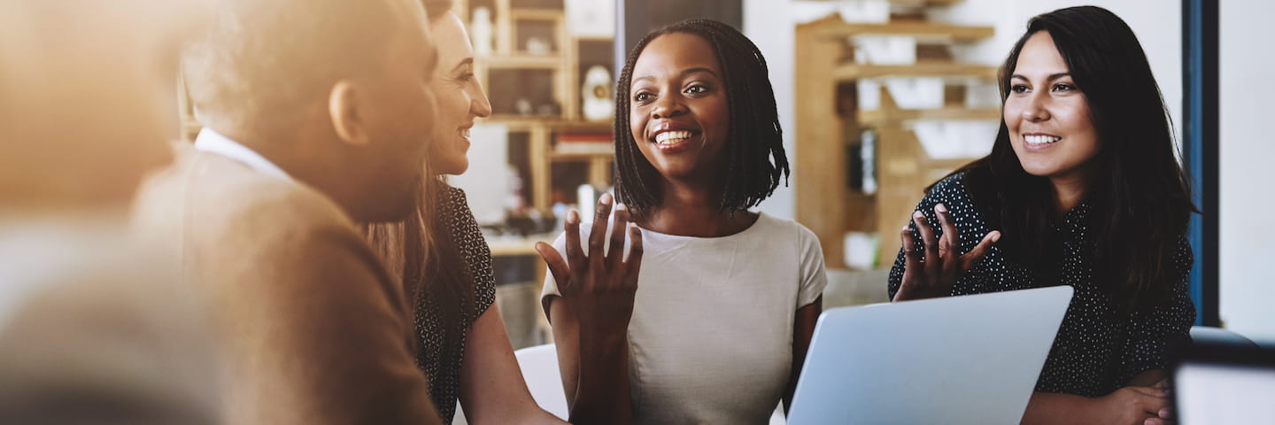 Diverse group of business professionals with online business management degrees having a lively conversation around a table in a modern office, smiling and gesturing as they engage with each other.