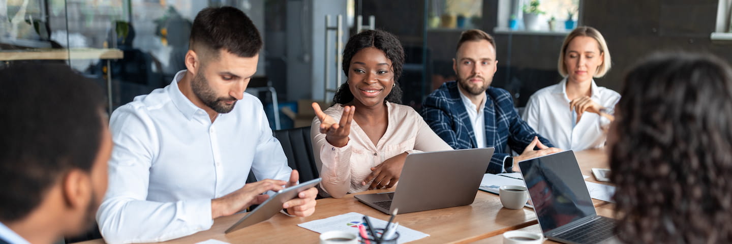 A diverse group of professionals is engaged in a business meeting at a conference table, discussing ideas with laptops, tablets, and papers about business degrees in front of them.