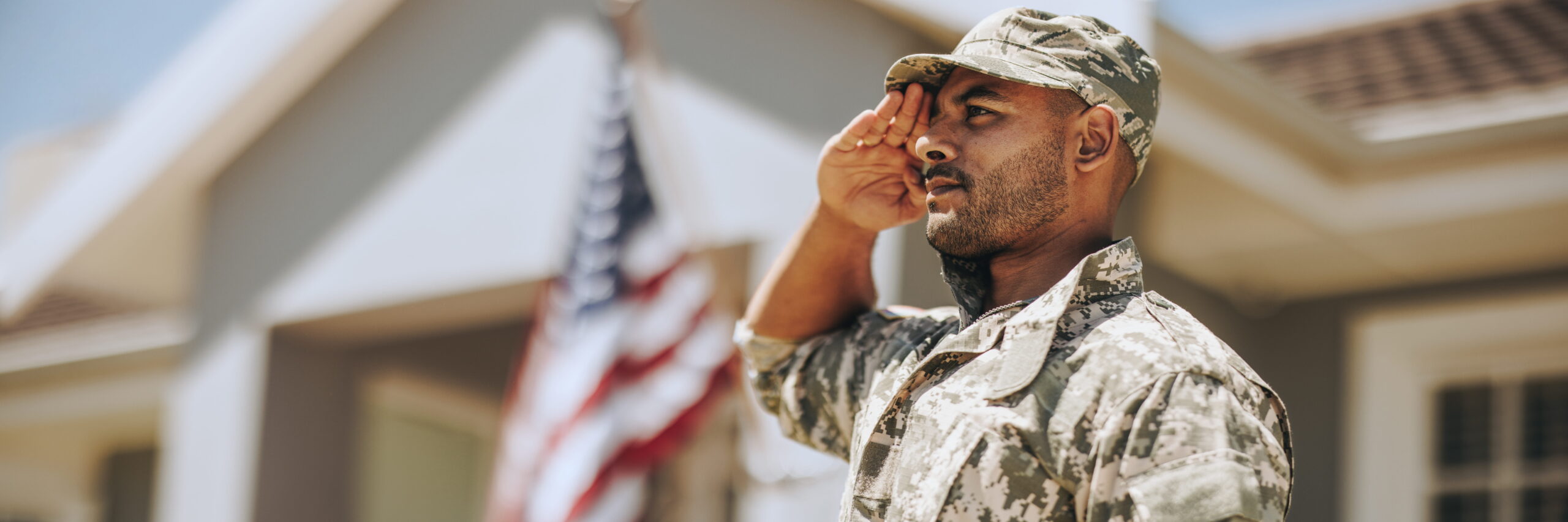 A male soldier in camouflage uniform saluting solemnly, with an American flag in the background against a clear blue sky outside a residential home, honoring veterans.
