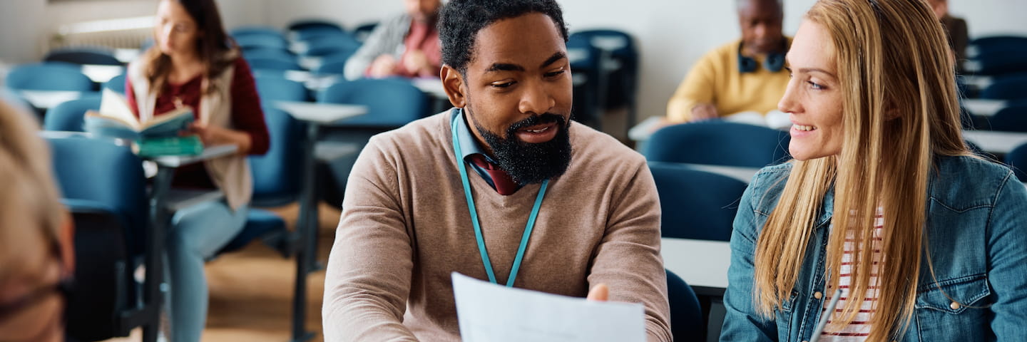 Two adult students, a man with a beard and a woman, are discussing over a paper in an online MBA classroom filled with diverse classmates.
