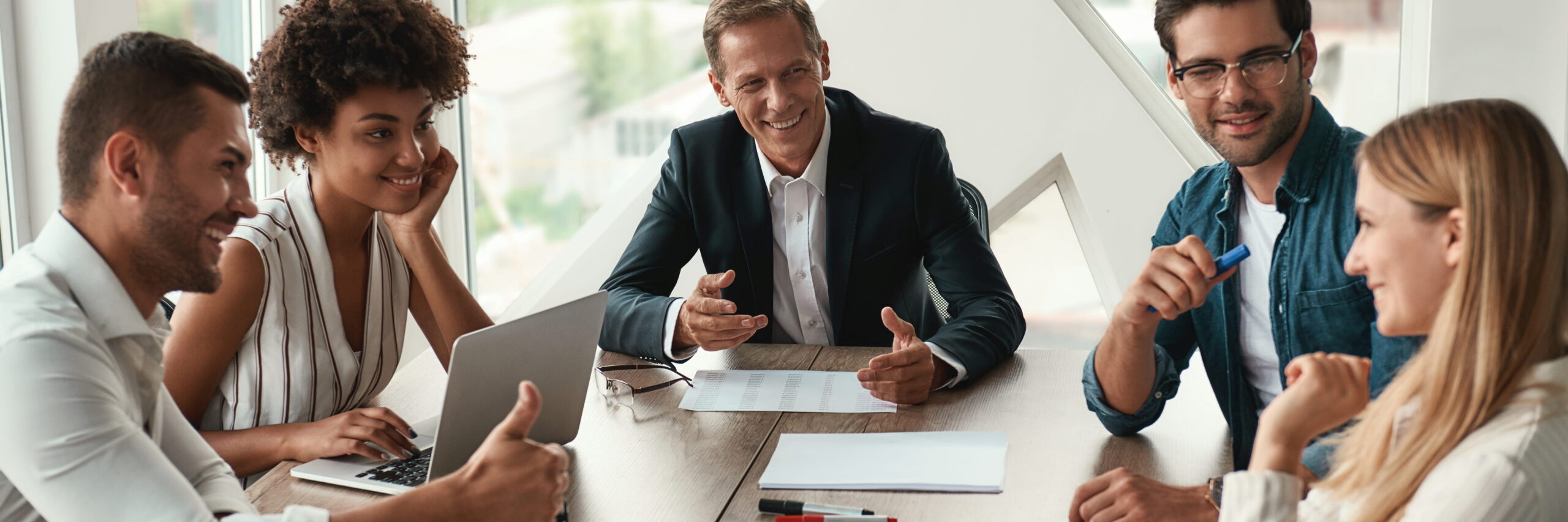 A group of five diverse adults in a business meeting, happily engaged in a discussion around a table with laptops and documents about accelerated online MBAs.