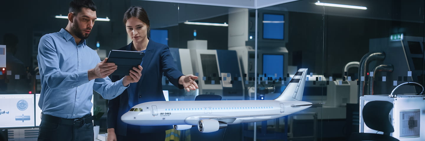 Two aerospace engineers, a man and a woman, discuss over a digital tablet next to a scale model of an airplane in a high-tech engineering laboratory while considering aspects of their online bachelor's in aerospace engineering.