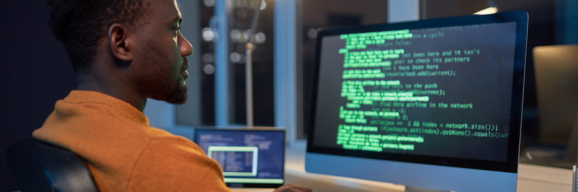 A man sits at a desk, focusing intently on a computer screen displaying lines of colorful code, in a dimly lit room as part of his online computer programming studies, with another device visible beside him.