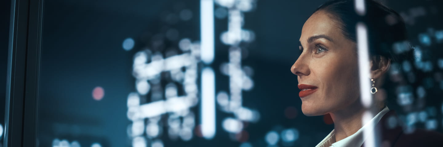 A professional woman gazes thoughtfully out of a window at night, with illuminated city buildings reflected on the glass, contemplating the best online PhD business programs for 2024.