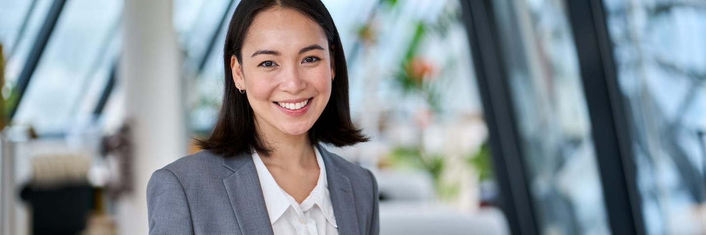 A professional portrait of a smiling Asian woman in business attire, standing in a modern office environment with large windows and green plants in the background, possibly contemplating her next step towards pursuing one of the best online business degrees.