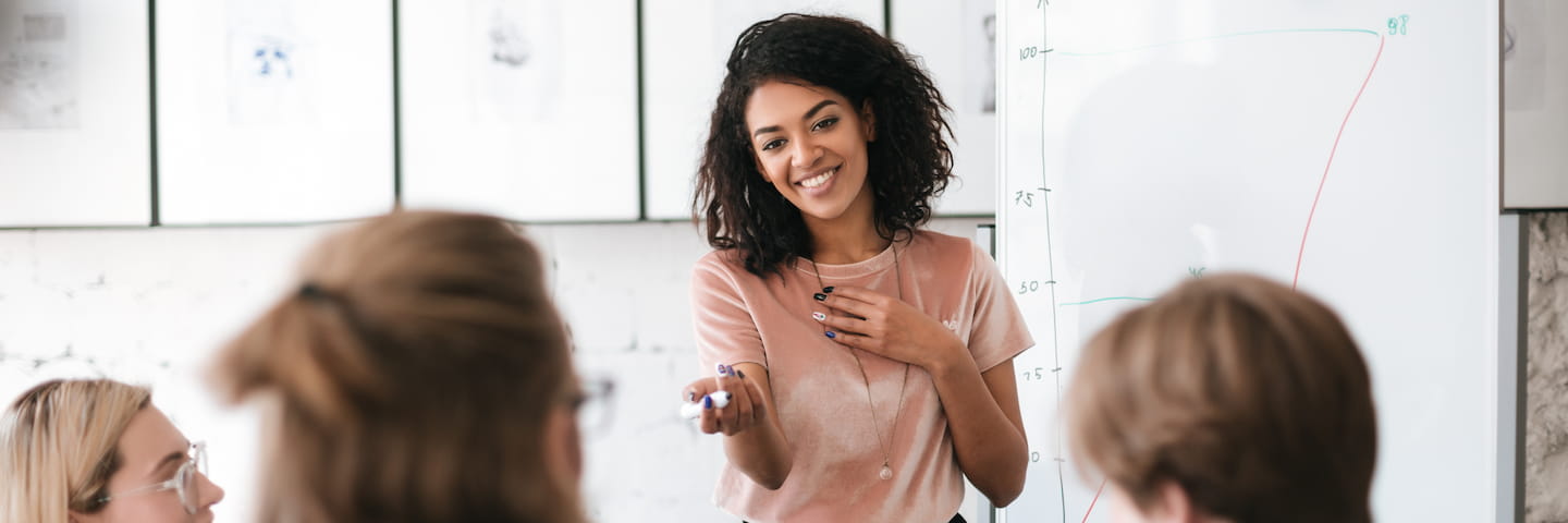 A smiling woman with curly hair presenting a graph on a whiteboard to a small group of attentive colleagues in a bright office setting, discussing the impact of online master's degrees on communications in 2024.