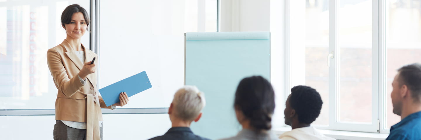 A professional woman in a beige suit holds a folder and presents communications strategies to a diverse audience in a bright conference room with a flip chart behind her.