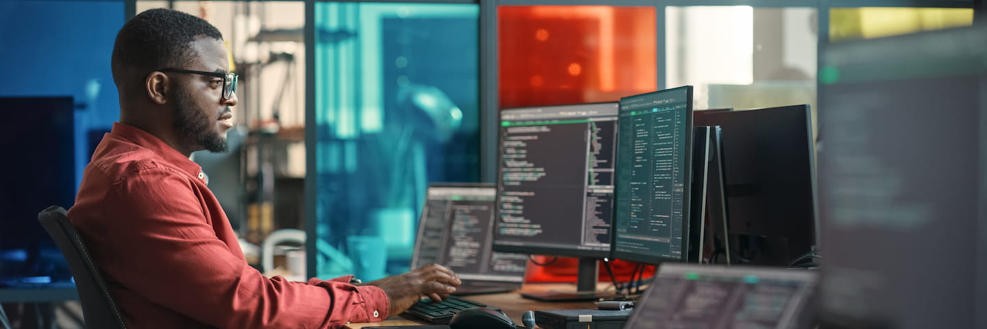 A focused man in a red shirt and glasses working at a multi-monitor workstation in a modern office environment, displaying coding and data analytics screens for his Computer Science Bachelor's Degree.