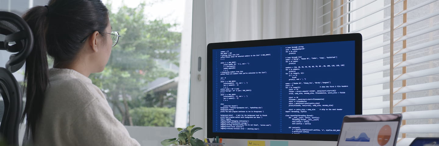 A woman in a wheelchair works on a computer displaying code, located by a window with blinds partially open, showing a view of trees outside. The screen also features tabs for researching the best online degrees in Computer Science.
