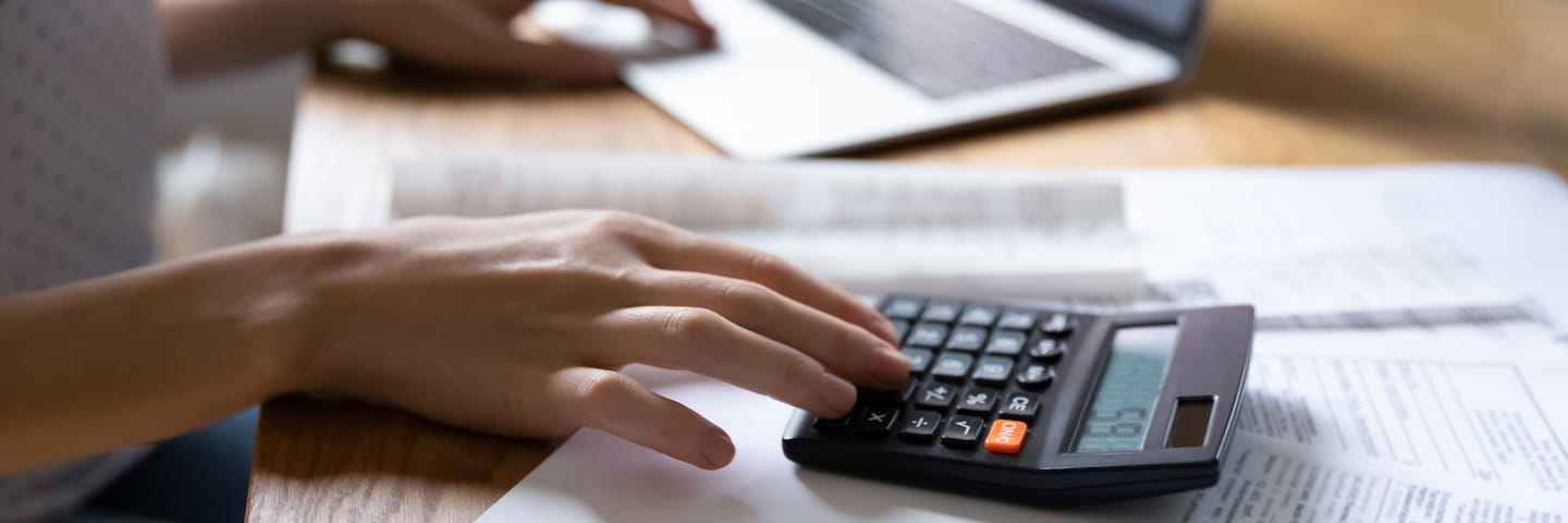 Close-up of a person's hand using a calculator next to a laptop on a wooden desk, implying calculations for online MBA costs or budget planning.