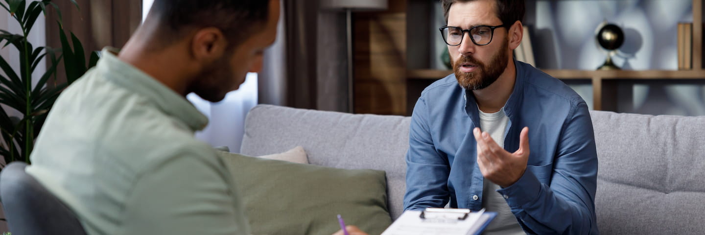 Two men engaged in a serious conversation about the best counseling degrees online. One man, wearing glasses and a blue shirt, gestures expressively while speaking. The other, in a green shirt, takes notes on a clipboard.