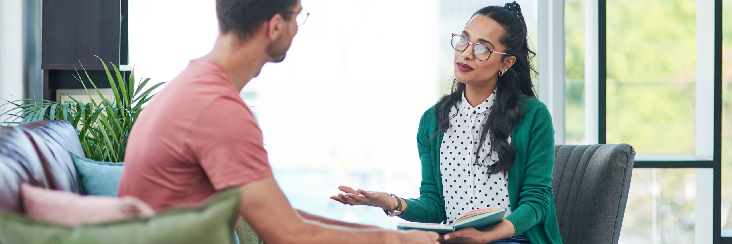 A professional counseling setting with a woman and a man sitting across from each other, engaged in conversation. The woman holds a notebook and listens attentively to the man. They are in a bright, modern office space.