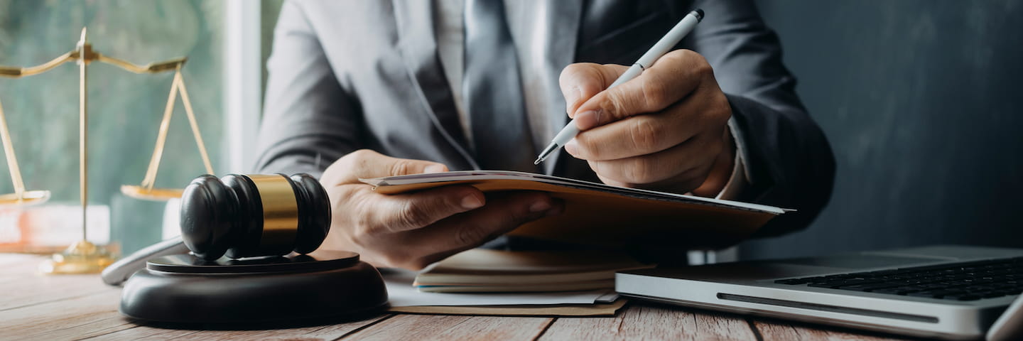 A lawyer in a business suit writes in a notebook at a desk with a gavel and scales of justice, symbolizing legal work. A laptop displaying information on online criminal justice associate degrees is also visible on the desk.