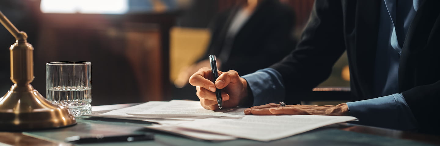 A person in a suit signing documents at a wooden desk with another individual partially visible, next to a bell and a glass of water, possibly reviewing materials for one of the best Criminal Justice degrees.